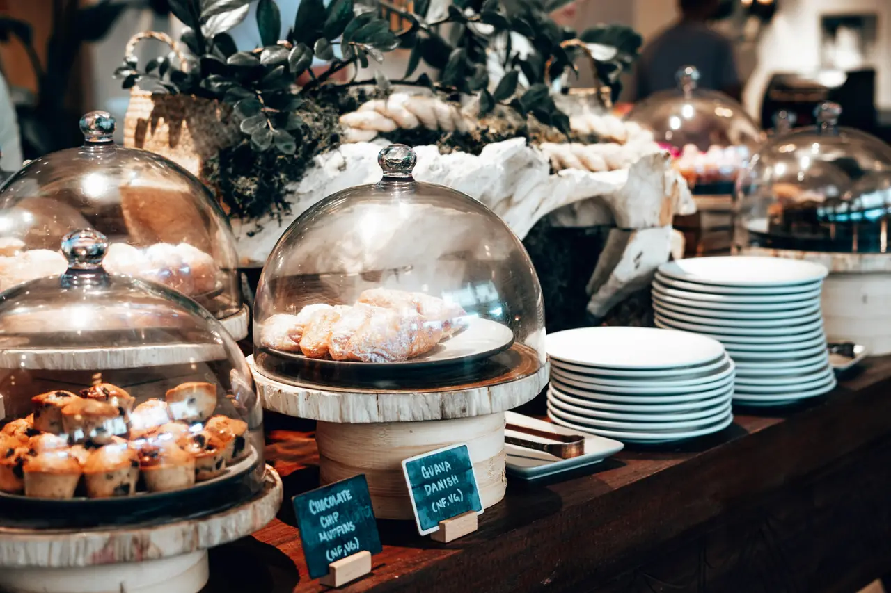 Mesón de buffet con la pastelería bajo campanas de vidrio, pilas de platos y fuentes calientes al fondo.