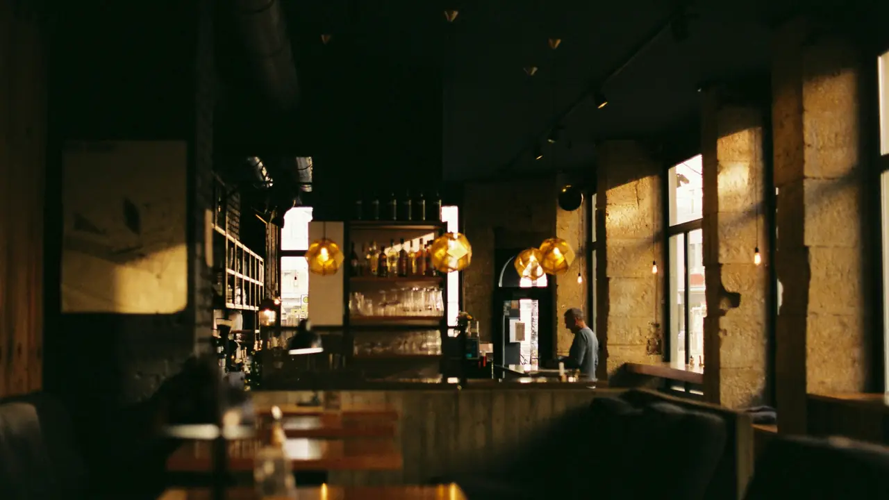 Salón en penumbra con la barra al fondo y ampolletas colgantes; la luz de la tarde entra por los ventanales y deja la mitad del cuadro en sombra.