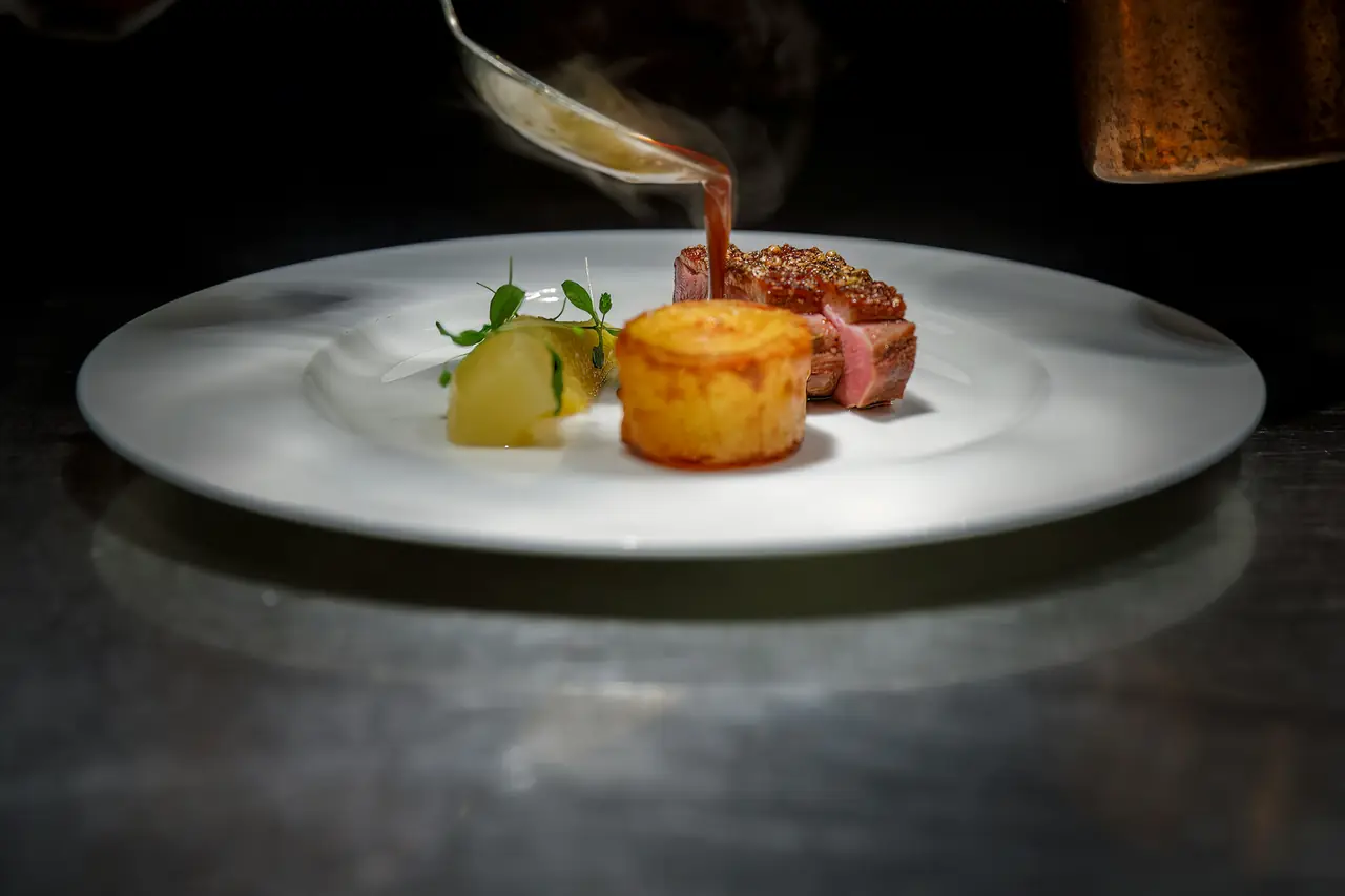 El cocinero termina el plato en el pase: vierte la salsa caliente sobre la carne ya montada.