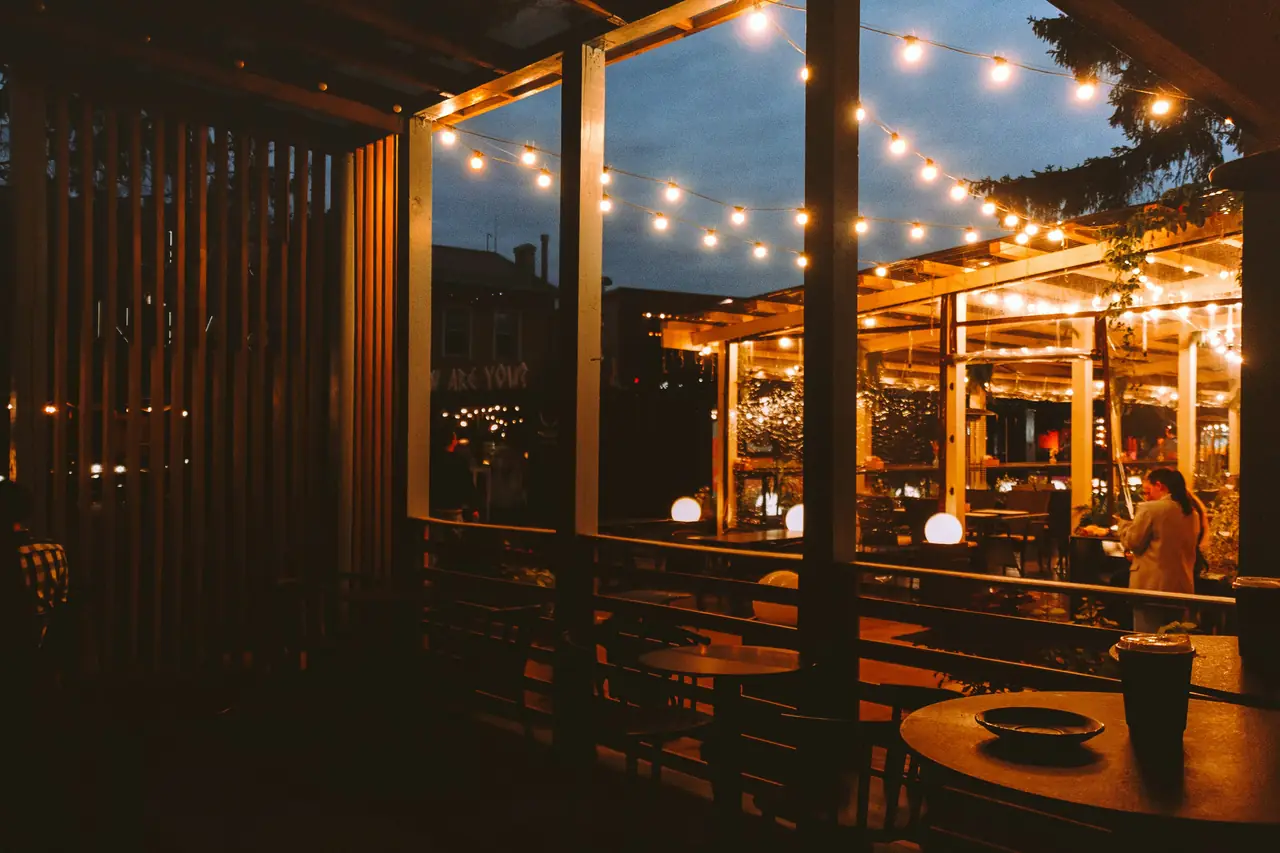 Terraza al anochecer bajo guirnaldas de ampolletas, con el salón encendido detrás del vidrio.